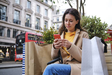 Junge Frau mit Einkaufstüten und Smartphone in der Stadt - ASGF00614