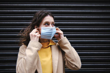 Woman looking away while wearing protective face mask in front of shutter during COVID-19 pandemic - ASGF00620