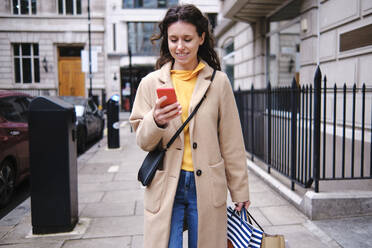 Smiling woman using smart phone while shopping in city - ASGF00624