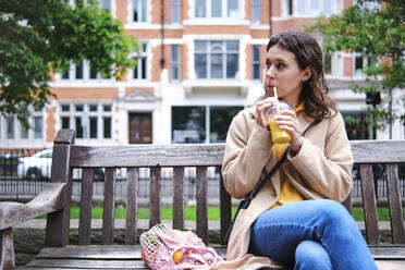 Young woman looking away while drinking smoothie on bench - ASGF00628