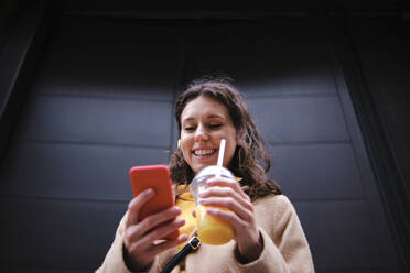 Smiling young woman with smoothie using smart phone near black door - ASGF00635