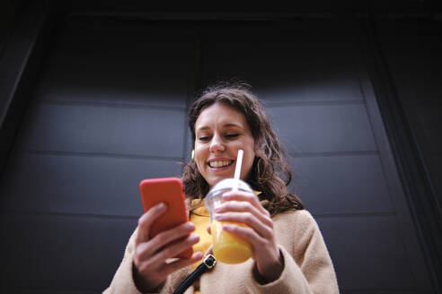 Lächelnde junge Frau mit Smoothie und Smartphone in der Nähe einer schwarzen Tür - ASGF00635