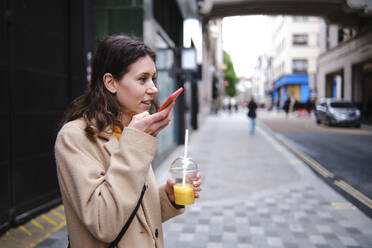 Junge Frau hält einen Smoothie in der Hand und sendet eine Sprachnachricht über ihr Smartphone auf dem Fußweg - ASGF00636