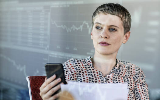 Weibliche Fachkraft, die ein Smartphone hält und eine Grafik auf einer digitalen Wand betrachtet - UUF23634