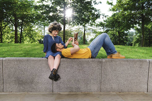 Boyfriend reading book while lying on girlfriend lap at park - ASGF00647