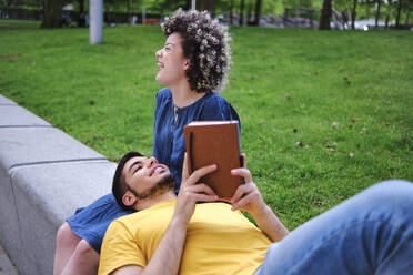 Junge Frau lacht, während ihr Freund mit einem Buch auf dem Schoß im Park liegt - ASGF00648