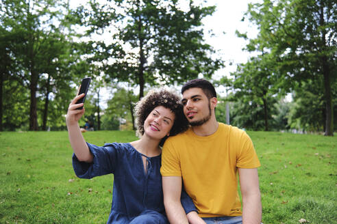 Lächelnde junge Frau, die ein Selfie mit ihrem Freund über ihr Smartphone macht, während sie im Park sitzt - ASGF00651