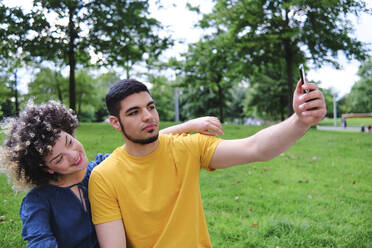 Junger Mann, der ein Selfie mit seiner Freundin über sein Smartphone macht, während er im Park sitzt - ASGF00653