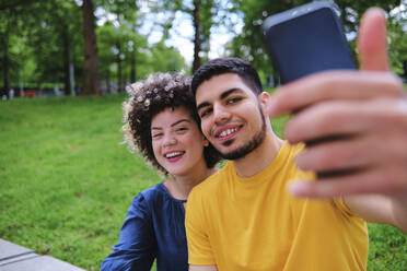 Glückliches junges Paar nimmt Selfie durch Smartphone sitzen im Park - ASGF00654