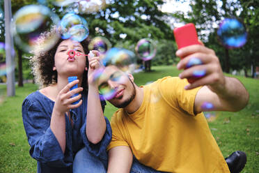 Junger Mann, der ein Selfie mit seiner Freundin macht, die im Park sitzt und Seifenblasen bläst - ASGF00668