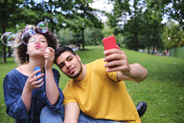 Junge Frau bläst Seifenblasen und sitzt neben ihrem Freund, der ein Selfie mit seinem Smartphone im Park macht - ASGF00669