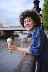 Beautiful happy curly haired woman with ice cream leaning on railing - ASGF00673