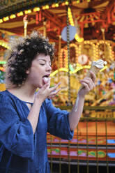 Young woman licking ice cream while standing at amusement park - ASGF00674