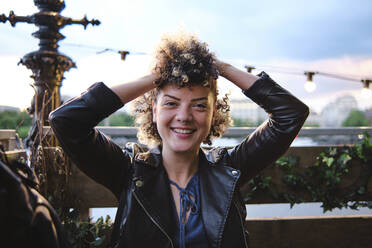 Happy beautiful young woman wearing leather jacket sitting with hand in hair - ASGF00675