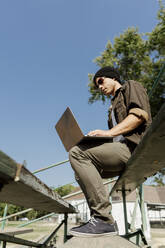 Junger Mann mit Sonnenbrille und Laptop auf einer Treppe im Park sitzend an einem sonnigen Tag - AFVF08972