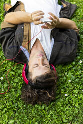 Young man using smart phone while listening music through headphones lying on grass - AFVF08973