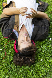Junger Mann, der sein Smartphone benutzt und über Kopfhörer Musik hört, während er im Gras liegt - AFVF08973