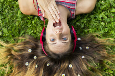 Shocked young woman listening music through headphones with flowers in hair lying on grass - AFVF08976