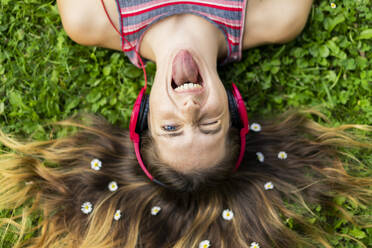 Schelmische junge Frau mit herausgestreckter Zunge und Blumen im Haar im Gras liegend - AFVF08977