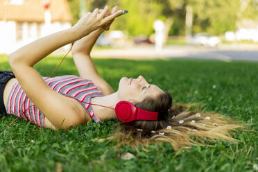 Young woman listening music through headphones using smart phone lying on grass - AFVF08979