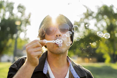 Junger Mann mit Sonnenbrille beim Blasen von Seifenblasen im Park - AFVF08980