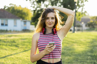 Beautiful young woman holding smart phone standing with hand in hair at park - AFVF08982