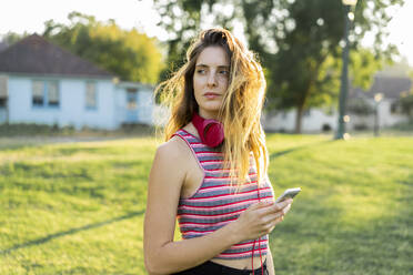 Beautiful young woman holding smart phone looking away standing at park - AFVF08983