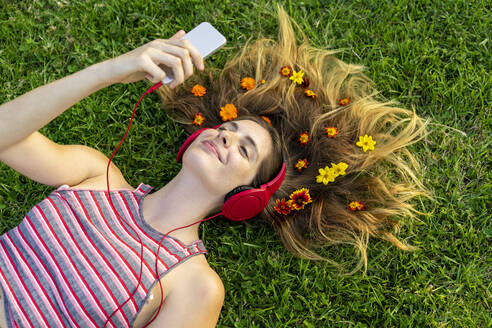 Lächelnde junge Frau mit Blumen im Haar liegt im Gras und hört Musik über Kopfhörer - AFVF08984