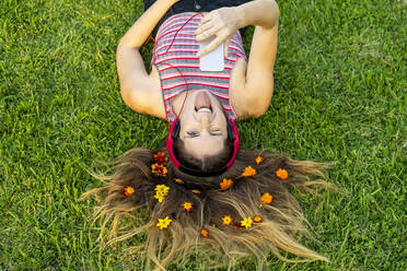 Mischievous young woman with flowers in hair sticking out tongue lying on grass - AFVF08985