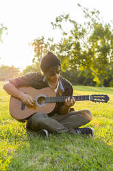 Junger Mann mit Mütze sitzt im Schneidersitz und spielt Gitarre im Park - AFVF08987