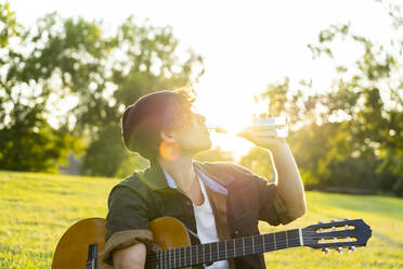 Junger Mann sitzt mit Gitarre im Freien und trinkt Wasser im Park - AFVF08991