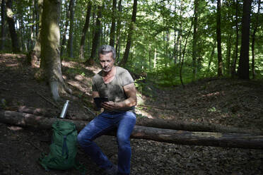 Male hiker using digital tablet while sitting on fallen tree - FMKF07192