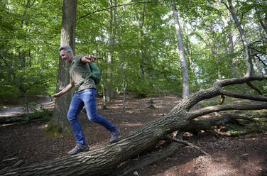 Unbekümmerter Mann mit ausgestreckten Armen, der auf einem umgestürzten Baum im Wald spazieren geht - FMKF07204