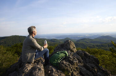 Älterer männlicher Wanderer mit Wasserflasche und Rucksack auf dem Gipfel eines Berges sitzend - FMKF07206