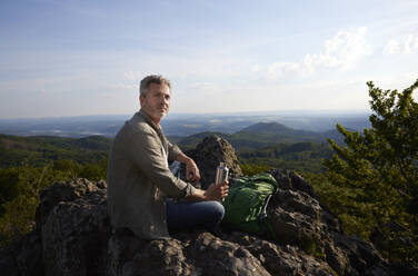 Männlicher Wanderer mit Rucksack und Wasserflasche auf dem Gipfel eines Berges sitzend - FMKF07207