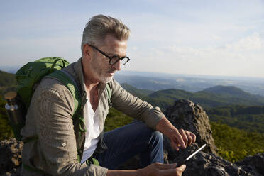 Männlicher Wanderer, der auf einem Berg sitzend ein digitales Tablet benutzt - FMKF07217