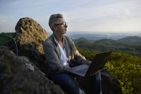 Mann hält Laptop und sitzt auf einem Berg - FMKF07237