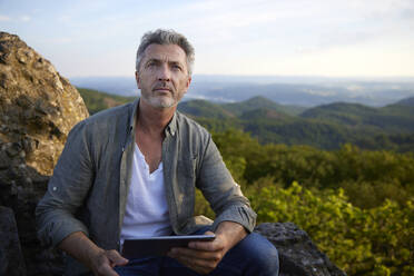 Männlicher Wanderer sitzt mit digitalem Tablet auf dem Gipfel eines Berges - FMKF07247