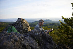 Männlicher Wanderer, der ein digitales Tablet benutzt, während er auf dem Gipfel eines Berges sitzt - FMKF07249