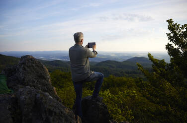 Mann fotografiert Landschaft durch digitale Tablet von Berg bei Sonnenuntergang - FMKF07250