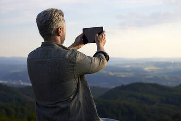Männlicher Wanderer, der bei Sonnenuntergang durch ein digitales Tablet fotografiert - FMKF07251