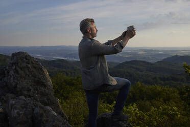 Männlicher Wanderer, der während des Sonnenuntergangs auf einem Berg durch ein digitales Tablet fotografiert - FMKF07252