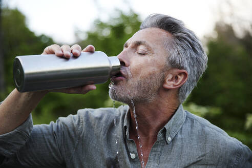 Thirsty man spilling while drinking water from bottle in forest - FMKF07265