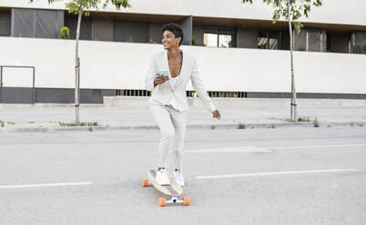 Happy female commuter holding smart phone while skateboarding on street - JCCMF02915
