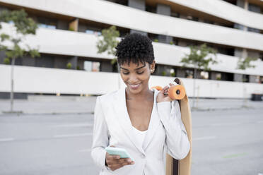 Lächelnde Geschäftsfrau, die ein Smartphone benutzt und ein Skateboard trägt - JCCMF02916