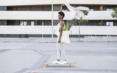 Businesswoman holding blazer while skateboarding with bag of vegetables on footpath - JCCMF02919