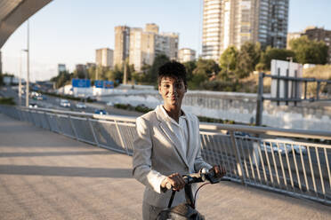 Businesswoman with electric scooter standing on footbridge in city - JCCMF02938