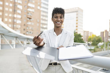 Smiling businesswoman offering agreement documents while standing on bridge - JCCMF02945