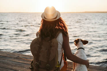 Woman with backpack and hat sitting by dog on pier - EBBF04077