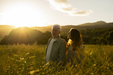 Smiling daughter looking at father while standing in field during sunset - AFVF09011